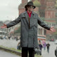 Person in a gray trench coat and hat standing on a city street with blurred background