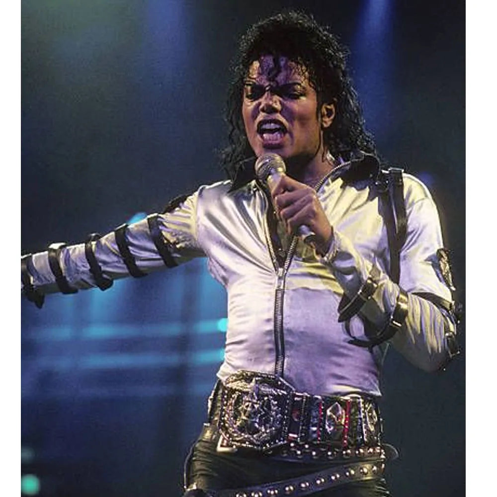 Person in a white jacket with black arm bands and a studded belt, holding a microphone on stage.