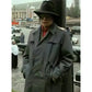 Person wearing a gray coat and hat in an outdoor setting with vehicles and buildings in the background.