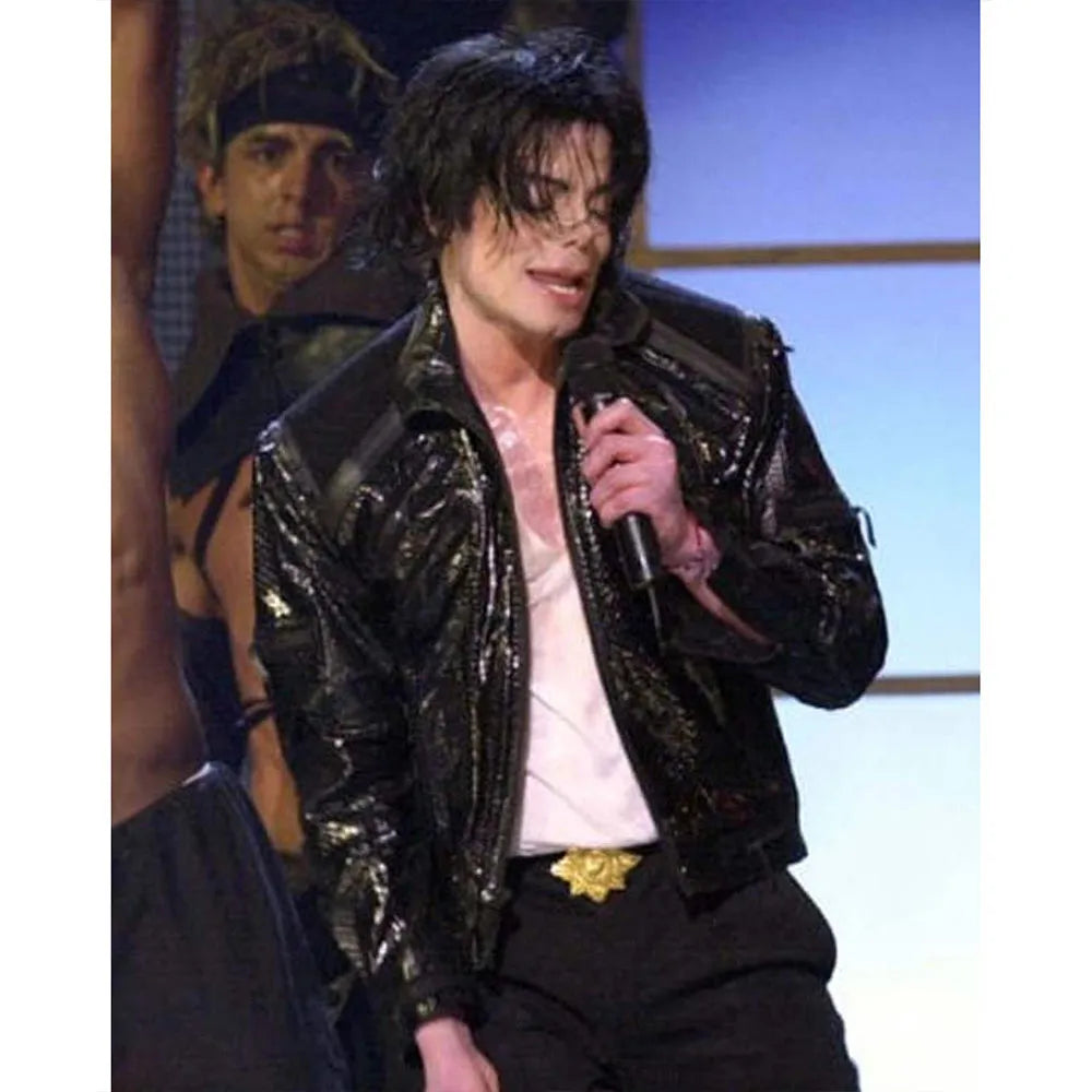 Person wearing a shiny black jacket on stage with another person in the background.